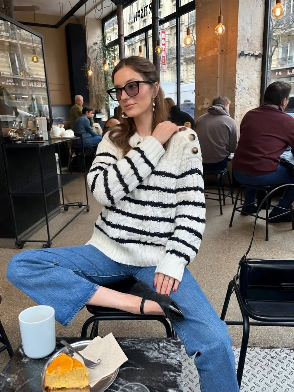 Woman sitting at a cafe table, posing with her hand on her shoulder, wearing a striped knit sweater and blue jeans.