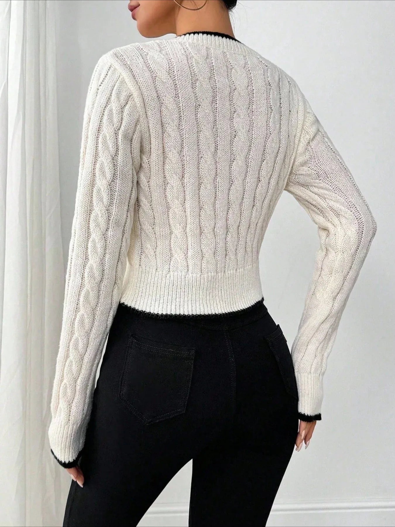 Back view of a woman wearing a white cable knit cropped V-neck sweater with black trim, paired with black pants.