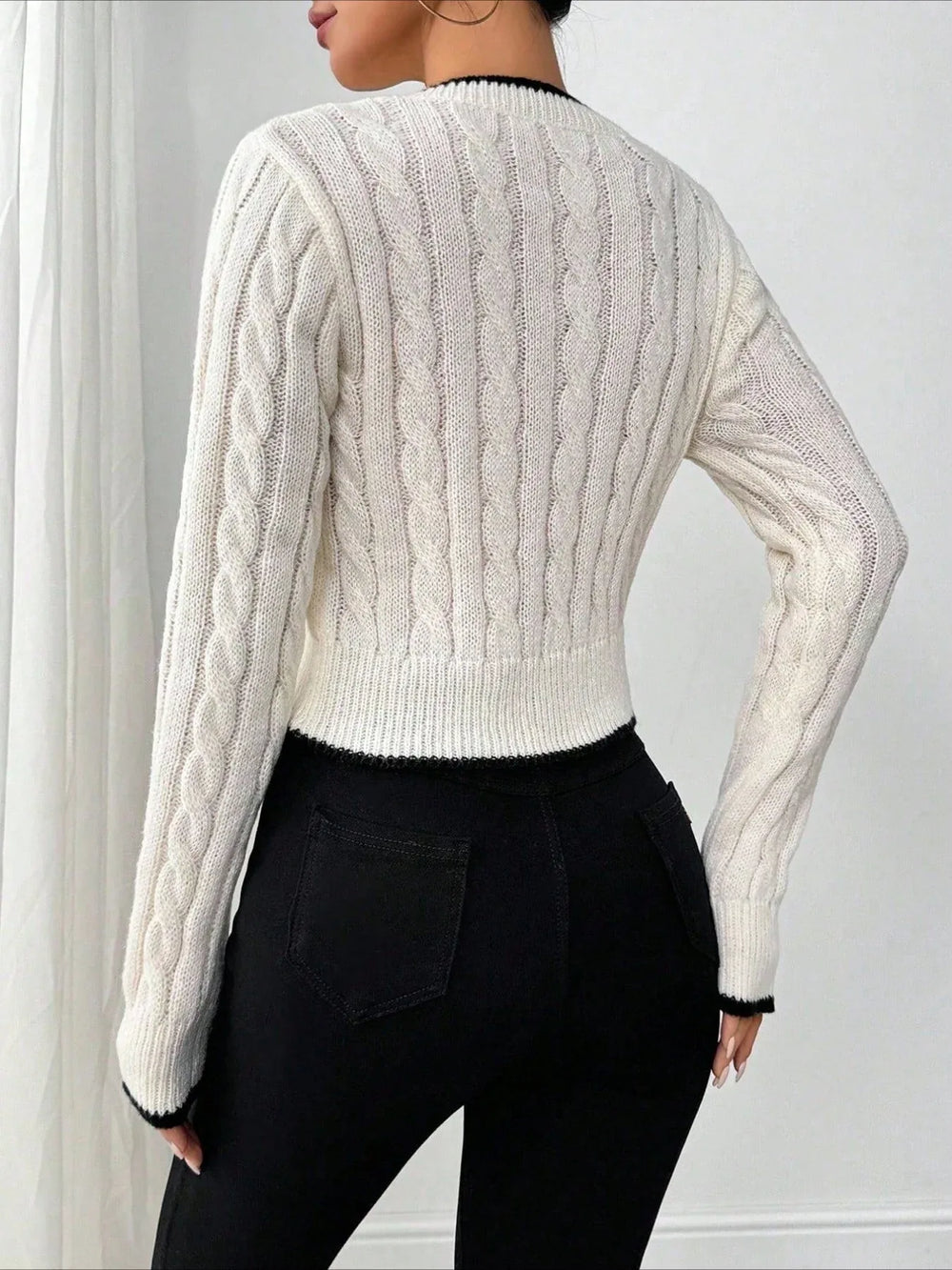 Back view of a woman wearing a white cable knit cropped V-neck sweater with black trim, paired with black pants.