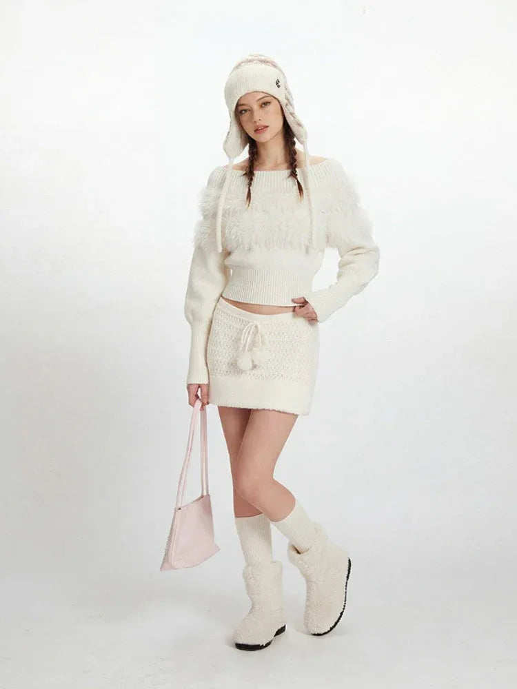 Full body shot of a model wearing a white off-the-shoulder sweater and matching mini skirt with a white knit hat, white knee-high socks, and white fuzzy boots. She is holding a pink handbag.