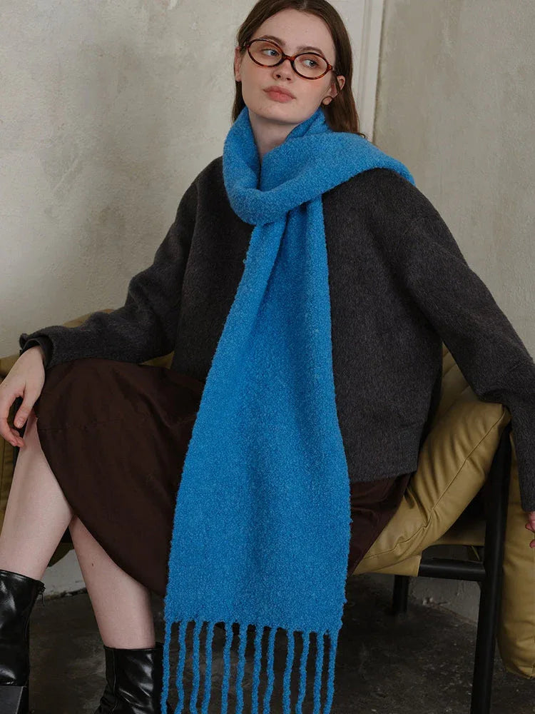 Woman sitting on a couch wearing a dark grey sweater and a bright blue textured knit scarf with fringe.