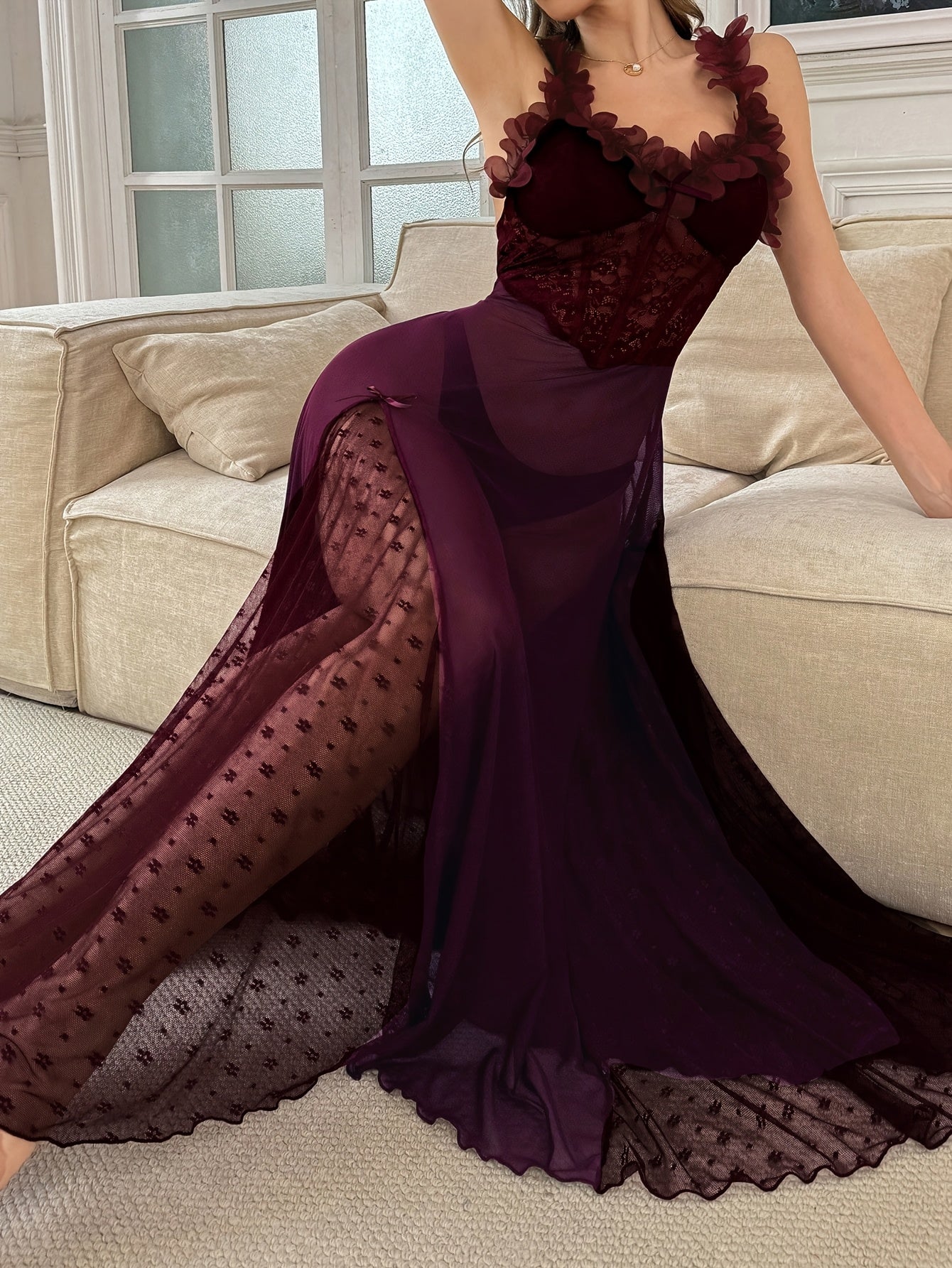Woman wearing a sheer plum maxi slip dress with floral applique straps and lace bodice, posing on a cream sofa.
