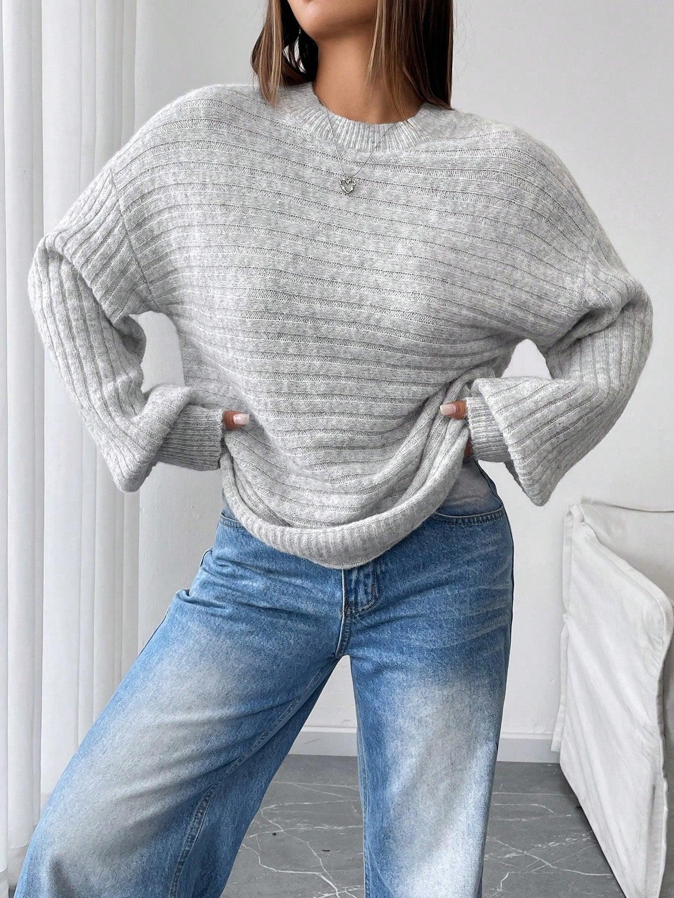 Woman posing with hands on hips wearing a light grey oversized knit sweater with balloon sleeves and a crew neck, paired with blue denim jeans.
