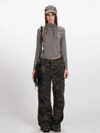 Woman wearing a grey long-sleeved mock neck top with a cropped hem and camouflage print wide-leg cargo pants, accessorized with a grey baseball cap and a black fringe crossbody bag, front view.