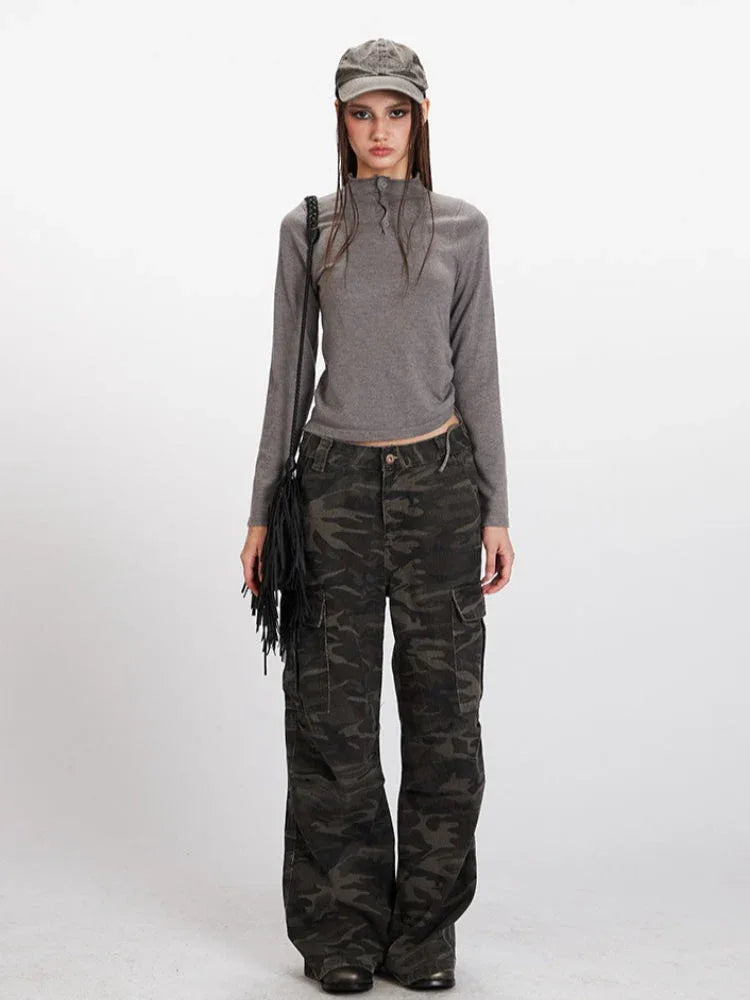 Woman wearing a grey long-sleeved mock neck top with a cropped hem and camouflage print wide-leg cargo pants, accessorized with a grey baseball cap and a black fringe crossbody bag, front view.
