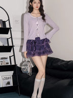 Full body shot of a woman wearing a lavender ribbed knit cardigan with lace trim and bow details over a white camisole, paired with a purple tiered ruffled skirt, white knee-high socks, and silver mary jane shoes, holding a silver handbag.