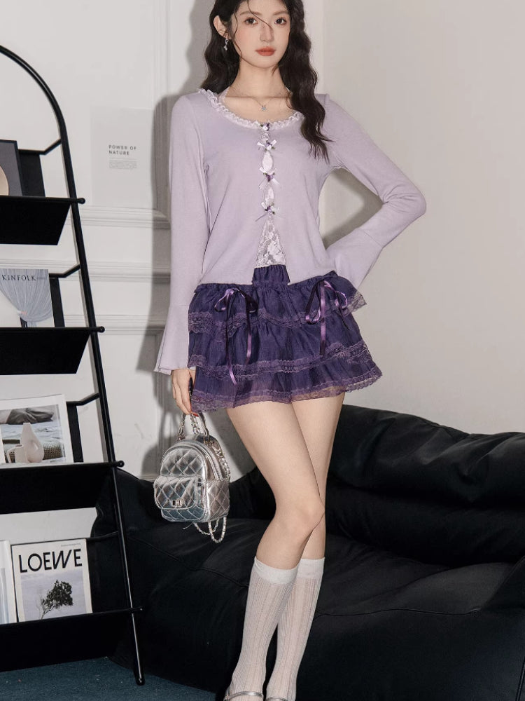 Full body shot of a woman wearing a lavender ribbed knit cardigan with lace trim and bow details over a white camisole, paired with a purple tiered ruffled skirt, white knee-high socks, and silver mary jane shoes, holding a silver handbag.