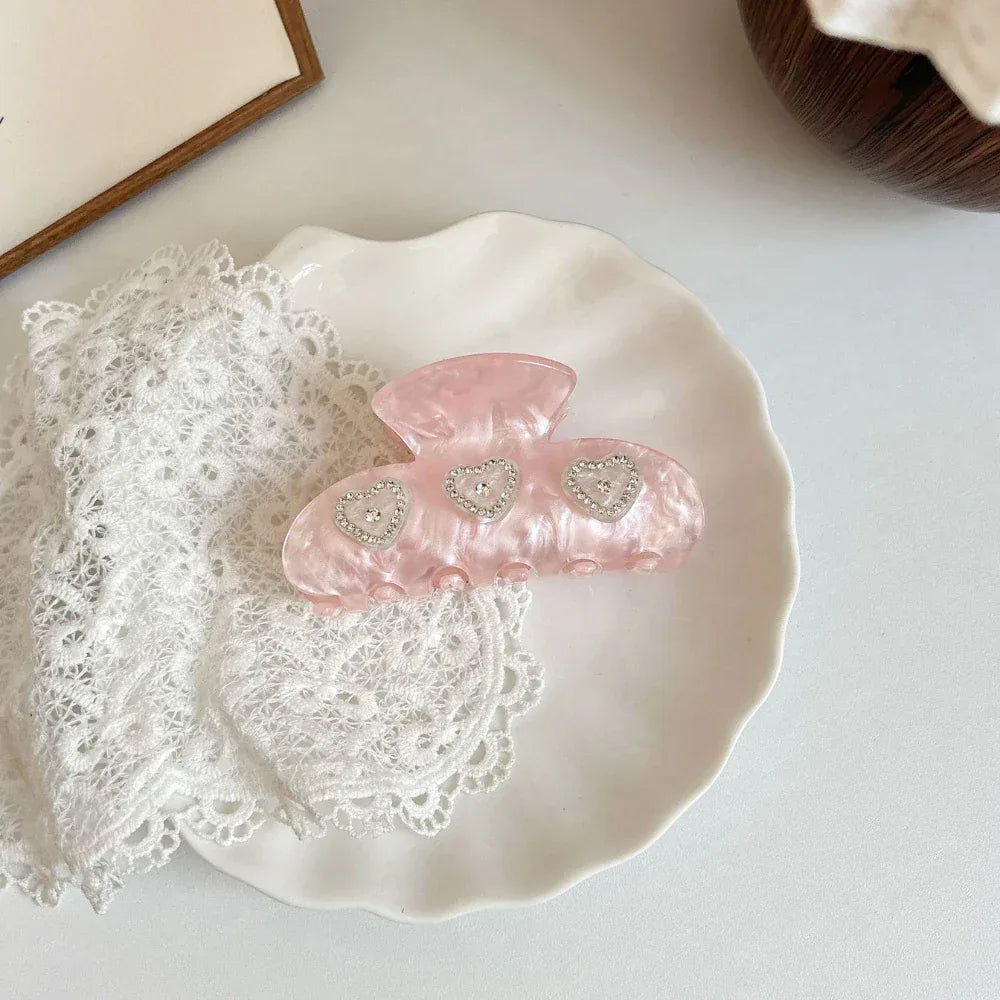 Pink marble claw clip with three heart-shaped embellishments adorned with rhinestones, placed on a white plate with white lace