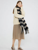 Full body shot of a woman wearing a cream cable knit sweater, a beige skirt, and a black and beige plaid scarf with fringe.