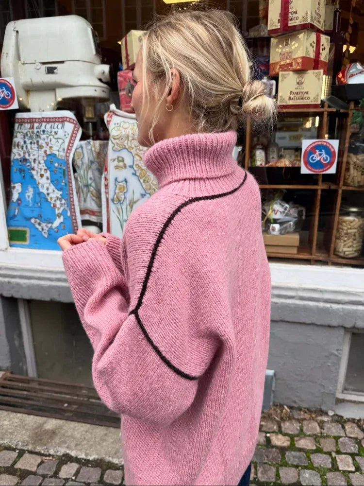 Side view of a woman wearing a pink oversized turtleneck sweater with contrasting black seam details and dark wash jeans.