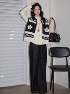 Full-body shot of a woman wearing a navy and cream floral and geometric knit vest over a cream long-sleeve top and dark wash wide-leg jeans, holding a handbag and striking a pose.