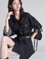 Woman wearing a dark wash denim shirt dress with a brown belt, sitting on a chair.