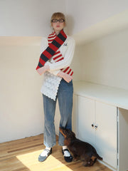 Model wearing a white sweater, blue jeans, and a red, grey, and white striped scarf, with a small dog.