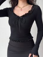 Woman wearing a black off-the-shoulder ribbed knit top with ruffled trim and tie front, paired with a black skirt and a heart pendant necklace.