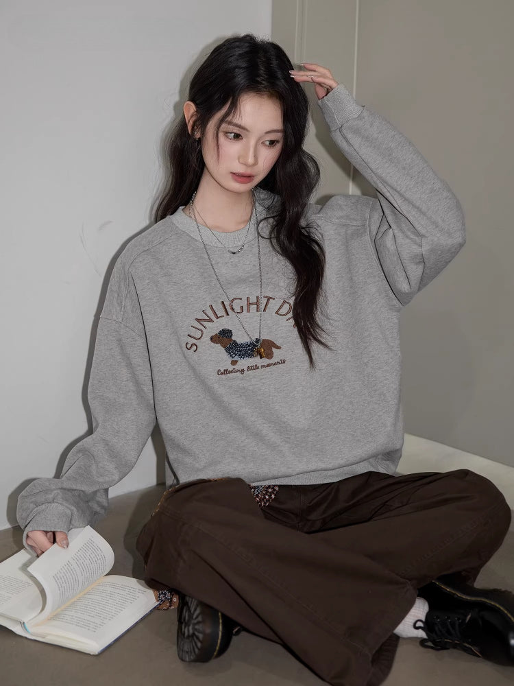 Woman sitting and reading a book, wearing a grey crew neck sweatshirt with a dachshund graphic and text "SUNLIGHT DAY".