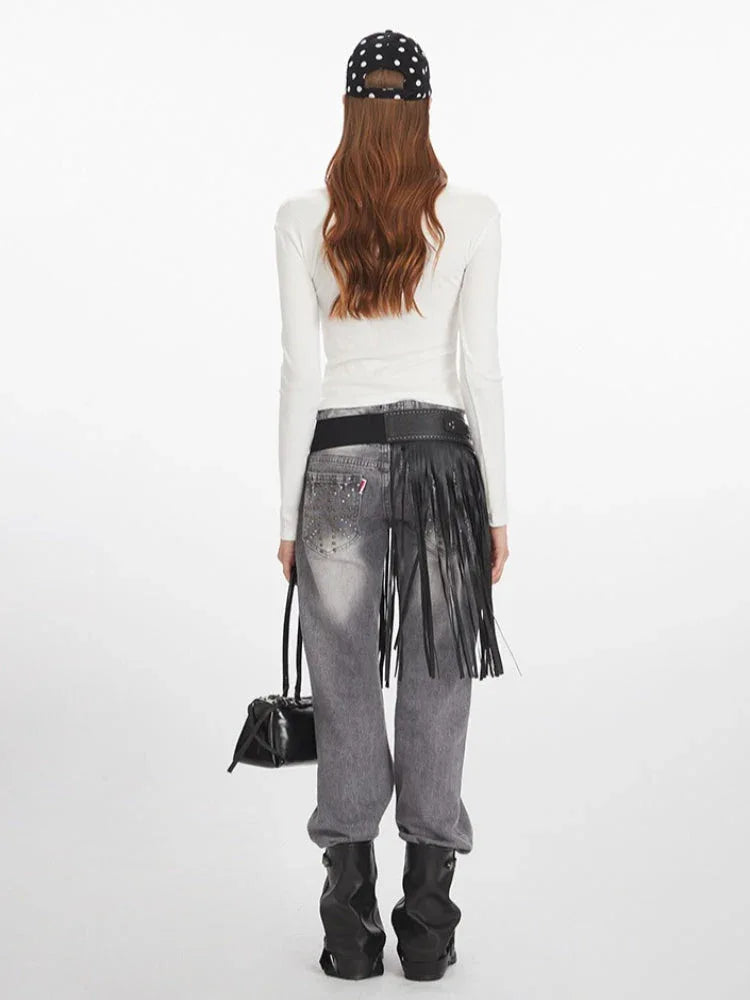 Back view of a woman wearing a white long-sleeved graphic t-shirt, grey distressed jeans with a black fringed belt, black boots, and holding a black handbag.
