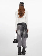 Back view of a woman wearing a white long-sleeved graphic t-shirt, grey distressed jeans with a black fringed belt, black boots, and holding a black handbag.