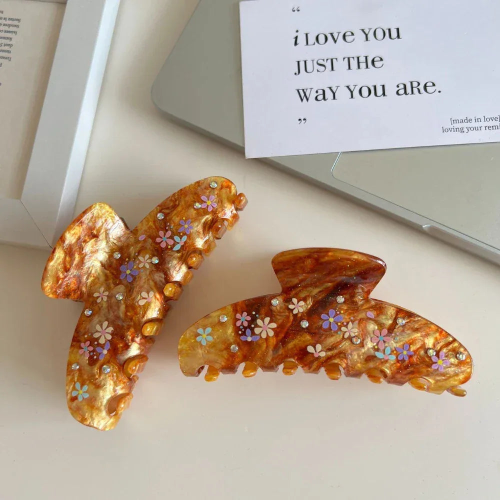 Two brown marbled hair claw clips with colorful floral patterns and gemstone accents are displayed on a white surface next to a laptop and a card.