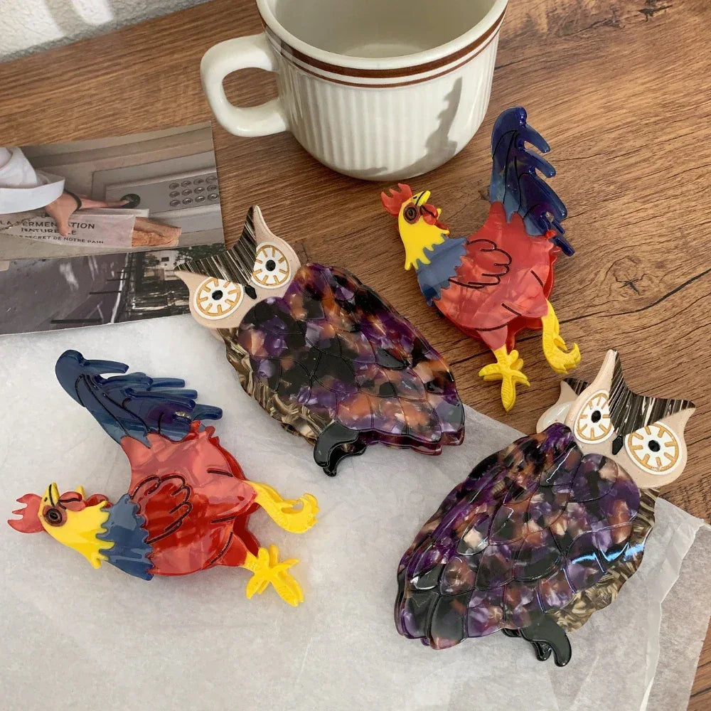 Four acrylic animal hair clips, two owls and two roosters, arranged on a wooden surface