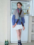 Model wearing a grey cropped sweater over a blue plaid long-sleeved shirt, white bubble skirt, and a colorful striped scarf, standing in front of an open refrigerator.