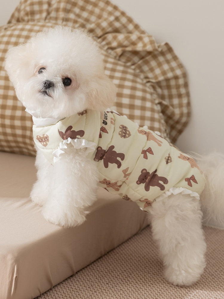 White poodle standing on a surface, wearing a light yellow pet vest with a teddy bear print and ruffle trim.
