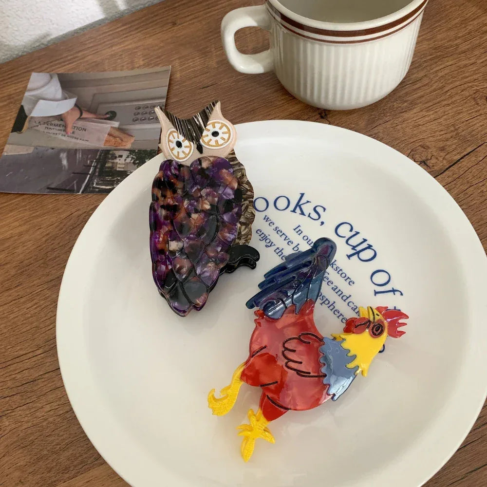 An acrylic owl hair clip and an acrylic rooster hair clip on a white plate
