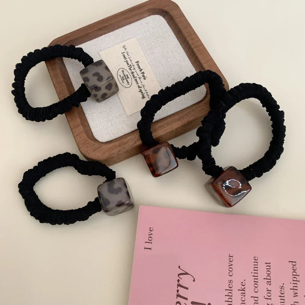 Three black velvet scrunchies with square leopard print pendants arranged on a wooden tray and a pink book