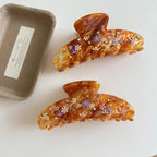 Two brown marbled hair claw clips with colorful floral patterns and gemstone accents are shown on a white surface next to a small cardboard container.