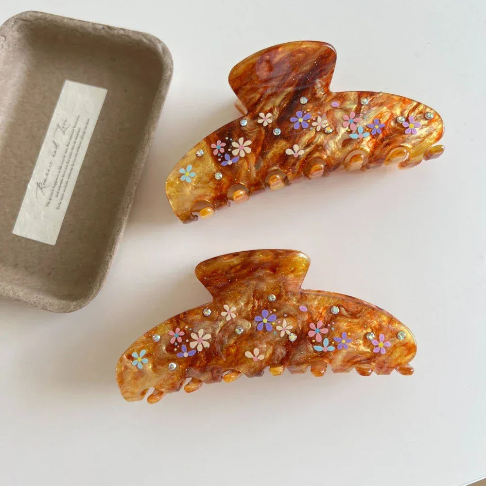 Two brown marbled hair claw clips with colorful floral patterns and gemstone accents are shown on a white surface next to a small cardboard container.