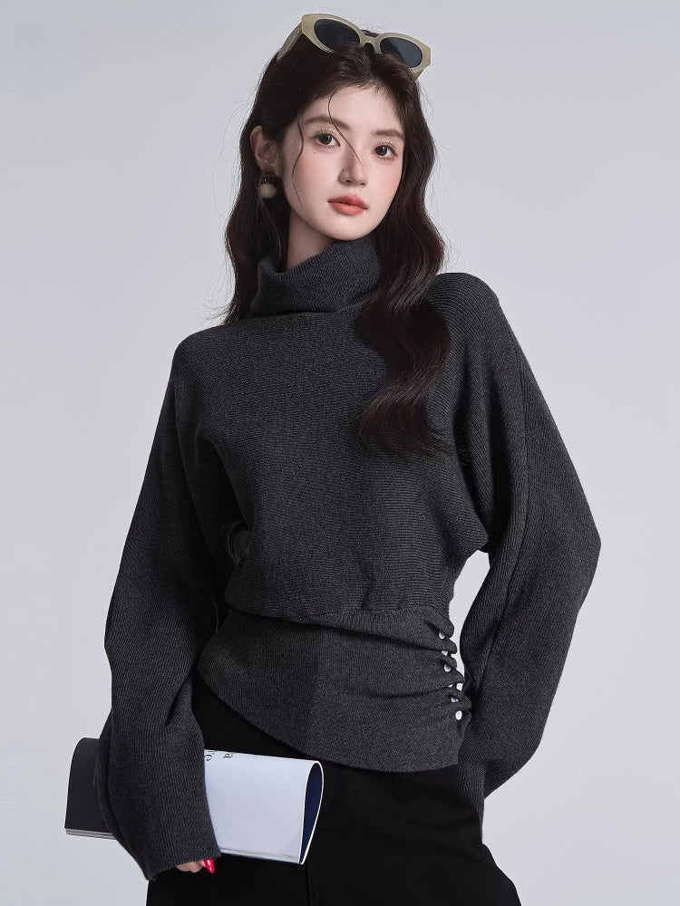 Close-up of a woman wearing a dark grey turtleneck sweater with batwing sleeves and a cinched button detail at the waist.