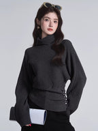 Close-up of a woman wearing a dark grey turtleneck sweater with batwing sleeves and a cinched button detail at the waist.