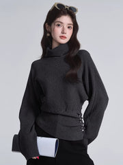 Close-up of a woman wearing a dark grey turtleneck sweater with batwing sleeves and a cinched button detail at the waist.