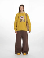 Woman wearing a yellow crew neck sweater with a bear graphic and brown wide-leg pants, with a brown crossbody bag.