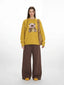 Woman wearing a yellow crew neck sweater with a bear graphic and brown wide-leg pants, with a brown crossbody bag.