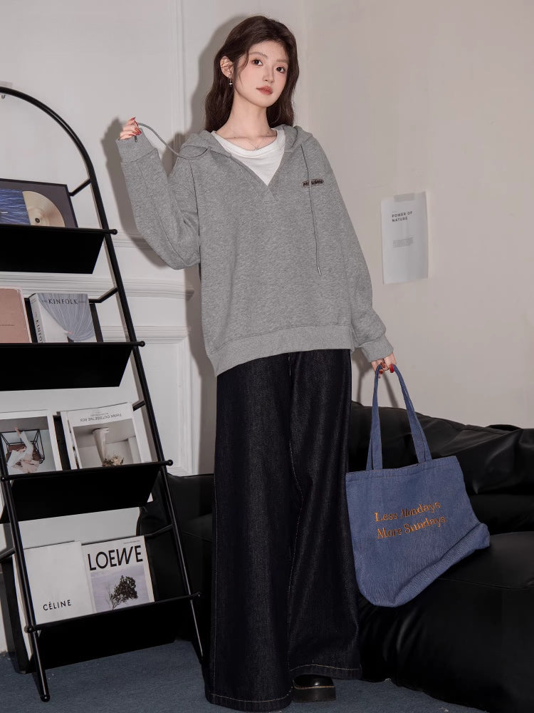 Full body shot of a woman in a grey V-neck oversized hoodie, black wide-leg jeans, and black shoes, holding a blue tote bag.