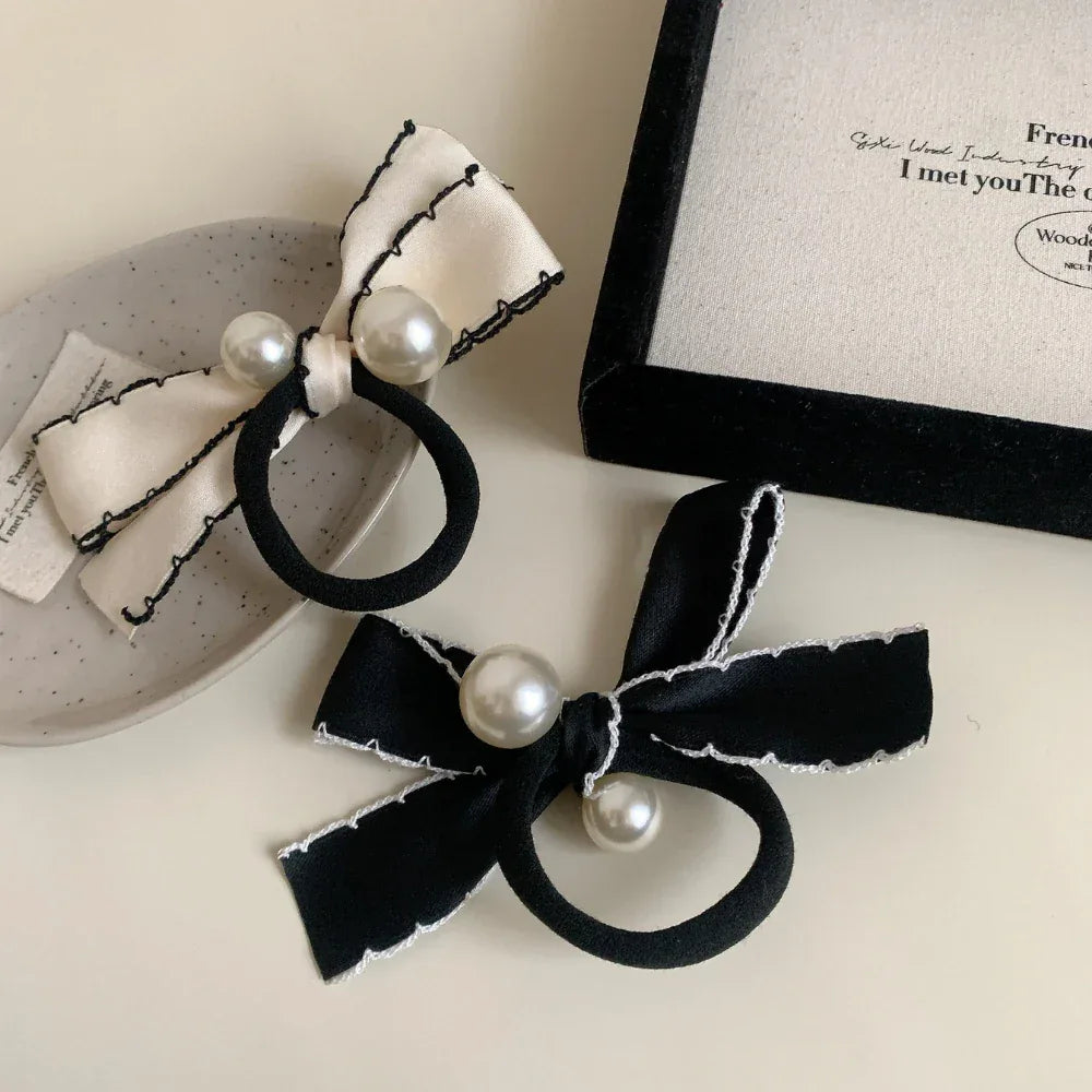 A cream bow hair tie with pearls is displayed on a speckled surface, and a black bow hair tie with pearls is positioned below it.