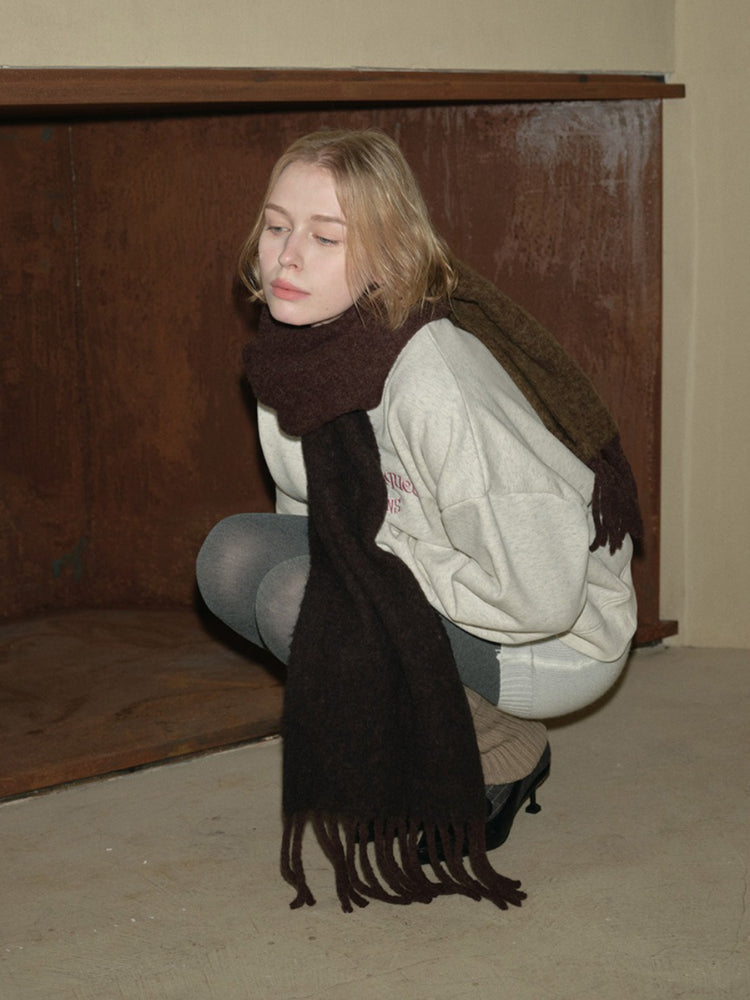 A young person wearing a cream-colored crew neck sweatshirt and a dark brown fringed scarf, looking down.