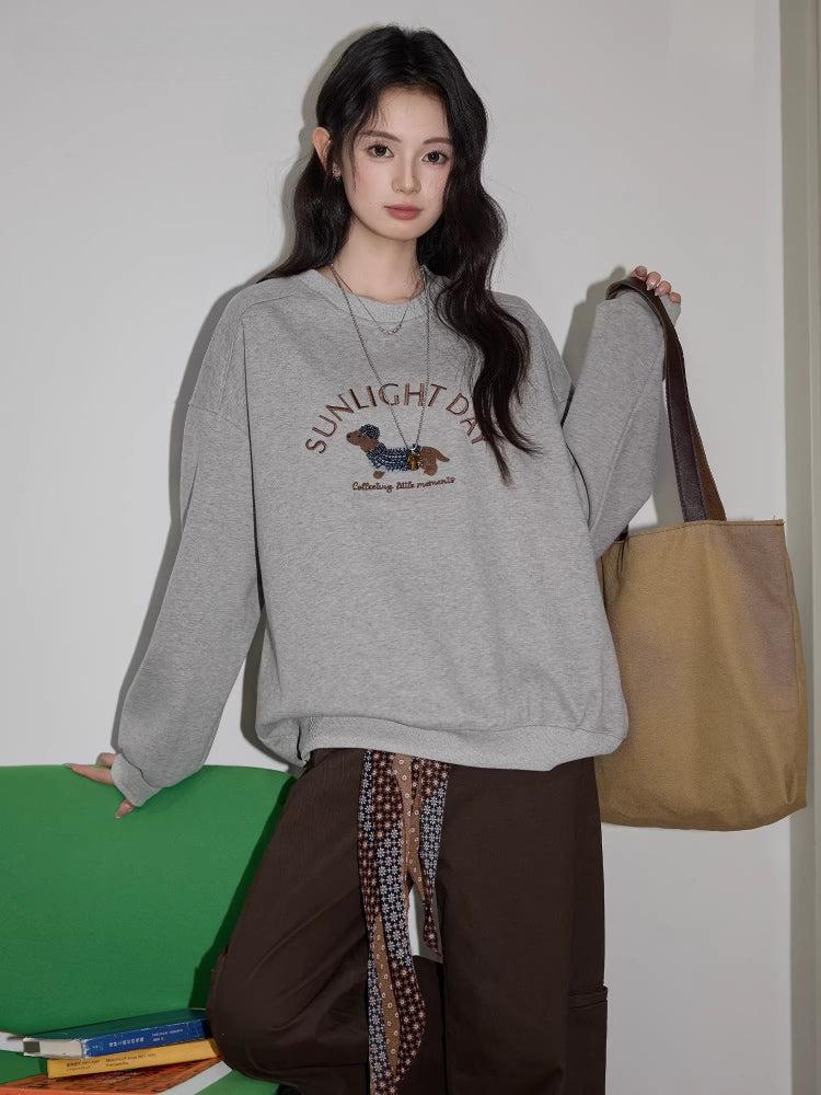 Woman wearing a grey crew neck sweatshirt with a dachshund graphic and text "SUNLIGHT DAY" on the front, paired with brown wide-leg pants and a tote bag.