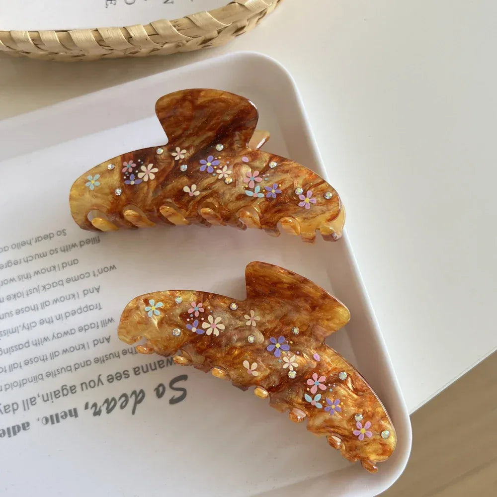Two brown marbled hair claw clips with colorful floral patterns and gemstone accents arranged on a white tray with text.