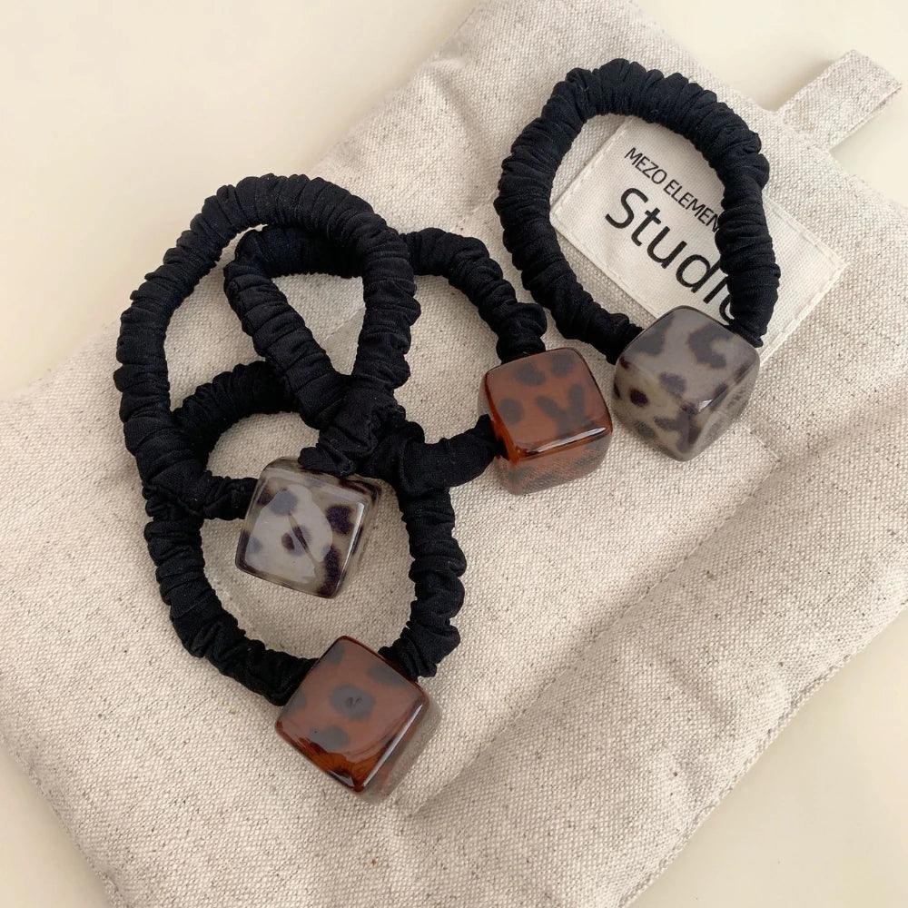 Group of black velvet scrunchies with square leopard print pendants displayed on a textured fabric surface