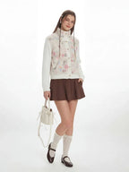 Full-body shot of a woman in a floral print fleece bomber jacket with cable knit sleeves, brown pleated skirt, white knee-high socks, and black Mary Jane shoes.