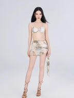 Woman in a sheer mesh abstract print bikini set with a ruched mini skirt and high-heeled sandals, standing against a white background.