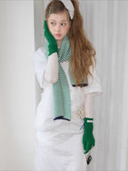 Model wearing a green and white striped knit scarf, white t-shirt, and green knit gloves with bow details, touching her face.