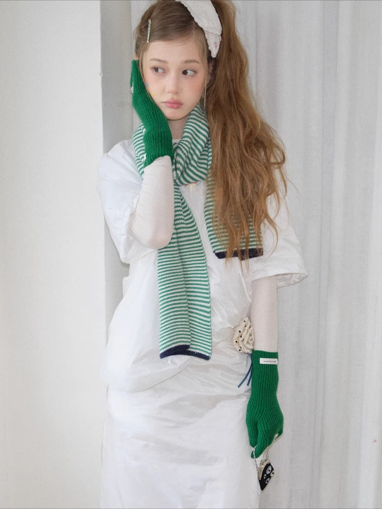 Model wearing a green and white striped knit scarf, white t-shirt, and green knit gloves with bow details, touching her face.
