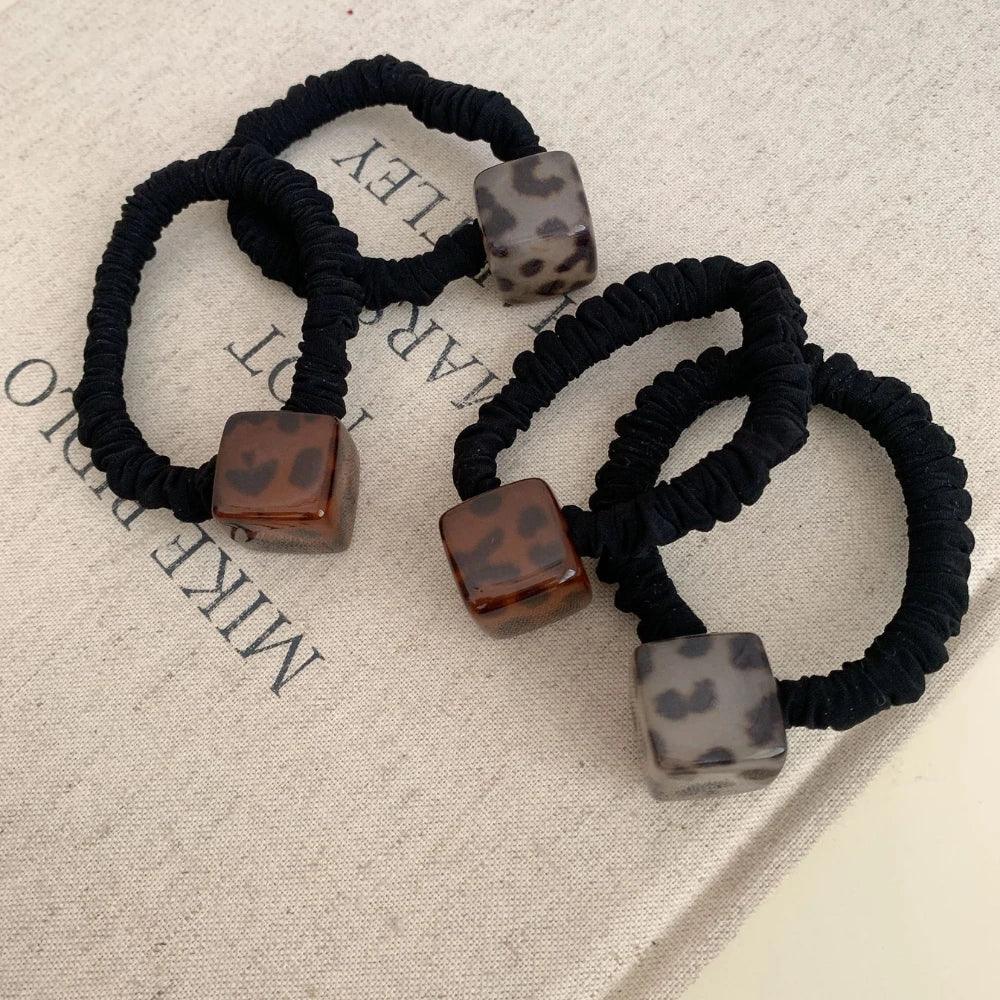 Close-up of three black velvet scrunchies with square leopard print pendants on a textured fabric background