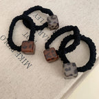 Close-up of three black velvet scrunchies with square leopard print pendants on a textured fabric background