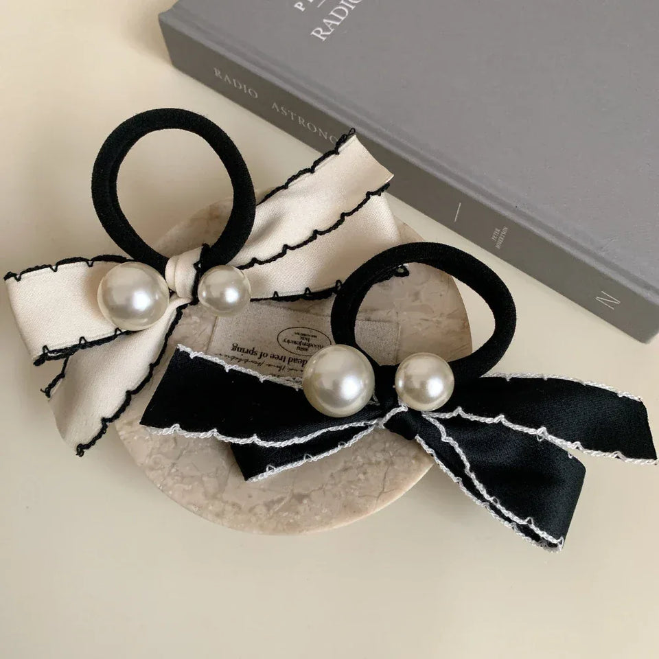 Two bow hair ties, one cream and one black, with pearl embellishments, are arranged on a surface with a book. Secondary image