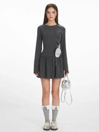 Woman wearing a dark grey long-sleeve mini dress with a gathered waist, white-trimmed grey socks, and silver shoes, holding a silver handbag and a small striped pouch around her neck.