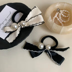 A cream-colored bow hair tie with pearl accents rests on a black decorative item, while a black bow hair tie with pearls sits nearby.