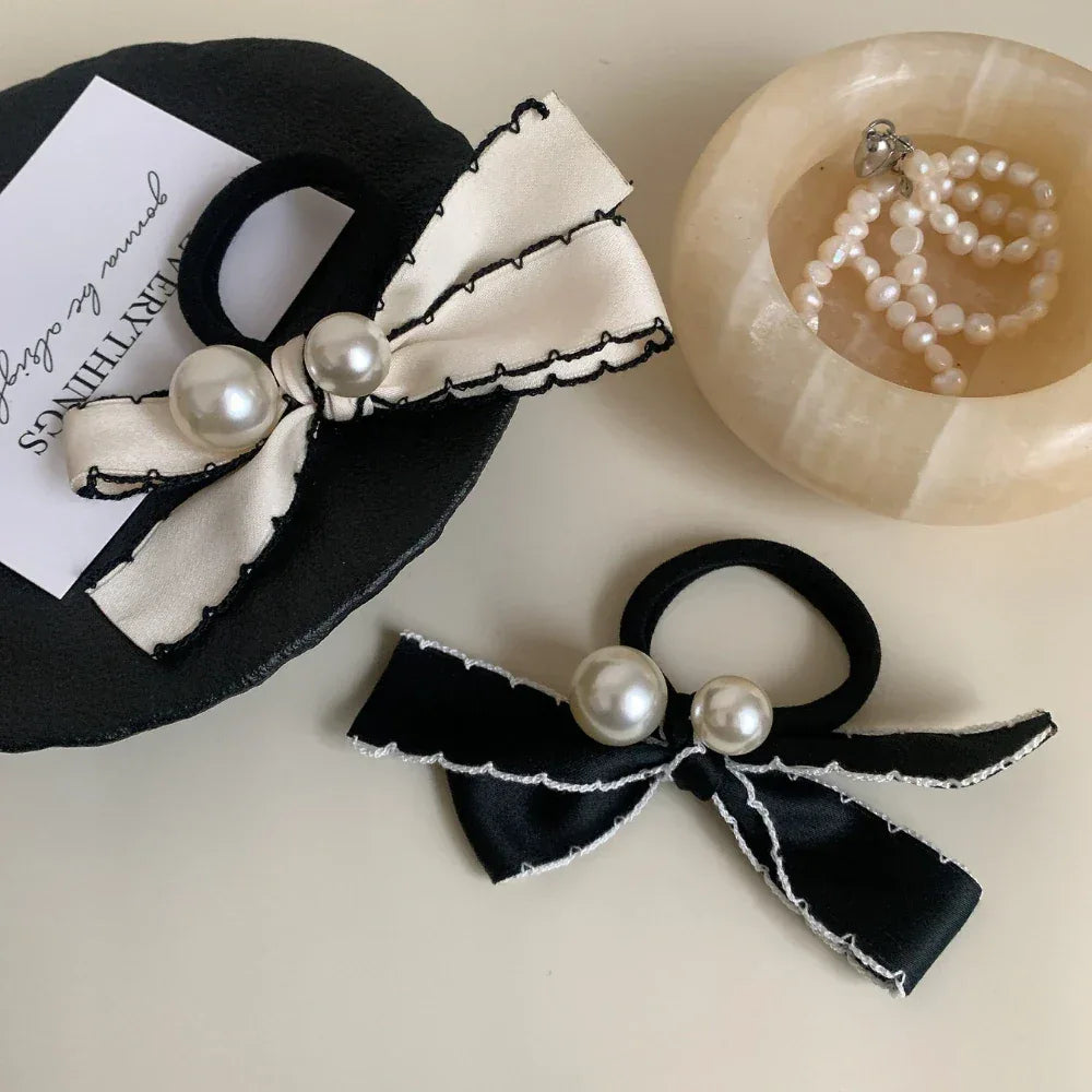 A cream-colored bow hair tie with pearl accents rests on a black decorative item, while a black bow hair tie with pearls sits nearby.